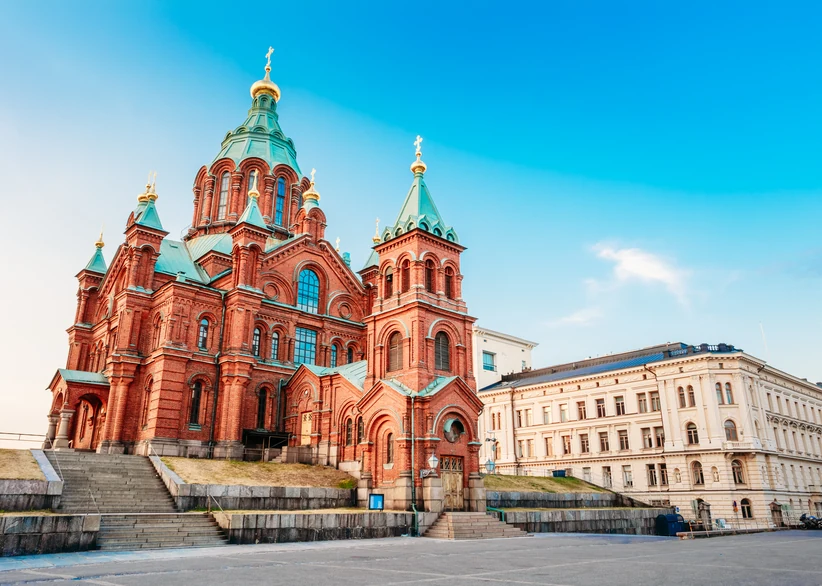 Uspenski Cathedral's red-brick facade and green onion domes topped with golden crosses rise above a stone staircase, with a white neoclassical building visible beside it under a bright blue sky.