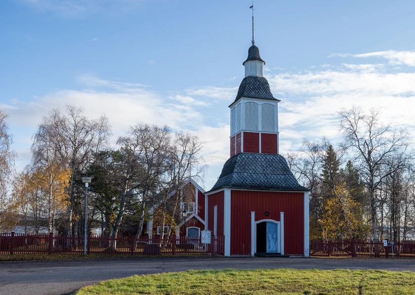 Jukkasjärvi Church's tall red and white wooden bell tower with its onion-shaped dome stands beside the red church building, surrounded by a red picket fence and bare autumn trees under a partly cloudy sky.