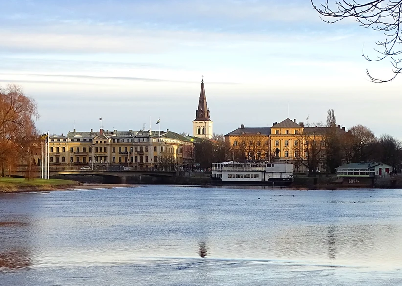 The town of Karlstad seen across a calm river, with a white church spire rising among historic yellow and cream buildings, a stone bridge, and a moored white boat along the waterfront under a hazy sky.