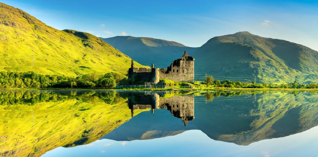 Kilchurn Castle ruins perfectly reflected in the still waters of Loch Awe with sunlit green hills and Highland mountains in the background, Argyll, Scotland