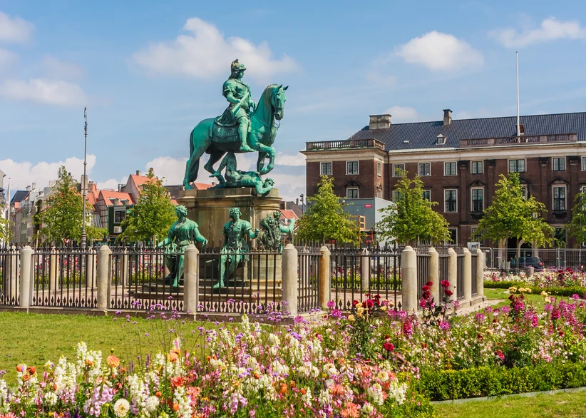 Green patinated bronze equestrian statue of King Christian V atop a fenced pedestal, surrounded by colorful flowerbeds, with historic townhouses lining the square in the background under a partly cloudy sky.