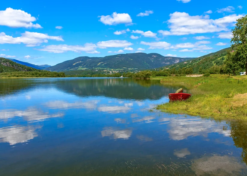 Lake Mjøsa mirrors a blue sky full of fluffy clouds, with a red rowboat resting on the grassy shore and forested mountains lining the far side of the water.