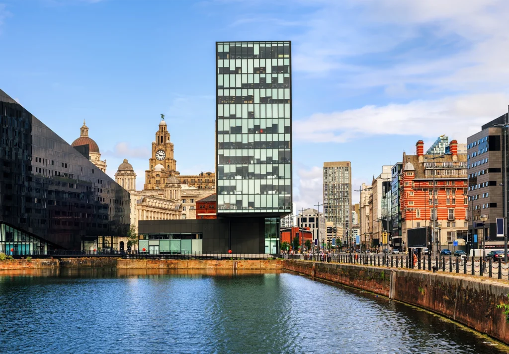 A glass tower and historic domed buildings line a calm dock basin under a bright blue sky.