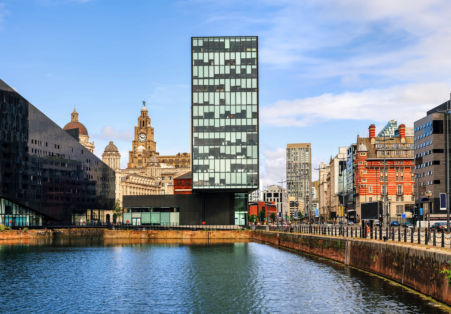 A glass tower and historic domed buildings line a calm dock basin under a bright blue sky.