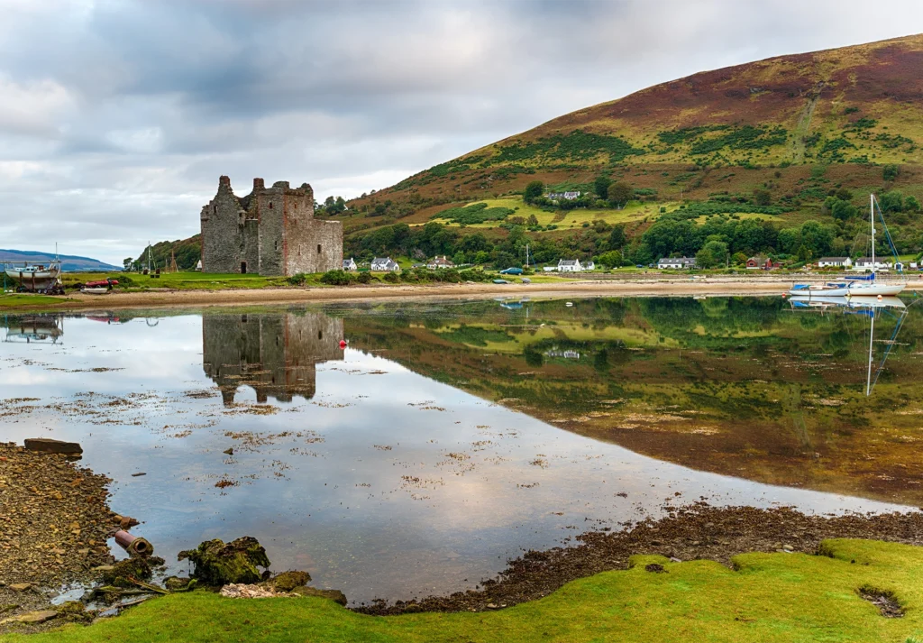 Ruined stone castle beside a calm bay, with moored yachts and a heather-covered hillside.