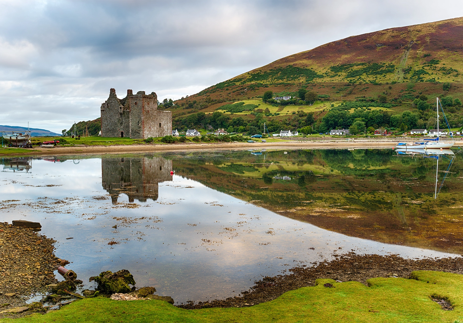 Ruined stone castle beside a calm bay, with moored yachts and a heather-covered hillside.