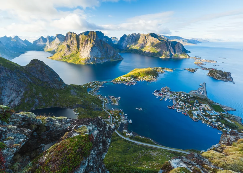 Panoramic view from Reinebringen peak overlooking the fishing village of Reine, with steep jagged mountains rising around a maze of blue fjord channels and scattered small islands under a bright sky.