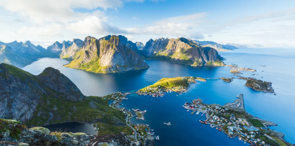 Panoramic view from Reinebringen peak overlooking the fishing village of Reine, with steep jagged mountains rising around a maze of blue fjord channels and scattered small islands under a bright sky.