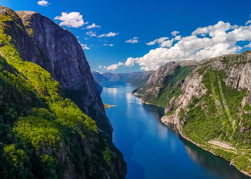 Aerial view down the length of Lysefjord, flanked by steep granite cliffs covered in patches of green vegetation, with the deep blue fjord winding into the distance beneath a partly cloudy sky.