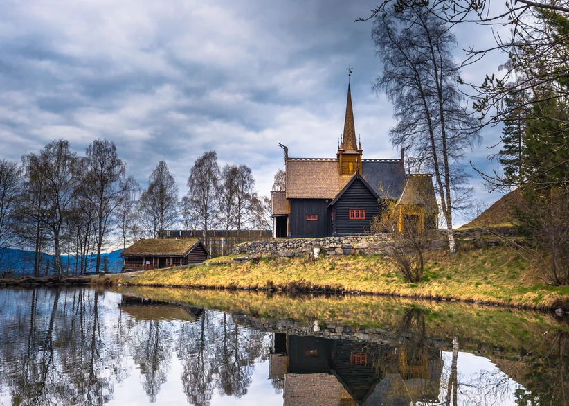 A dark wooden stave church with a tall spire and adjacent turf-roofed building sits on a grassy stone-walled bank, reflected in a still pond, at the Maihaugen open-air museum under a cloudy sky.
