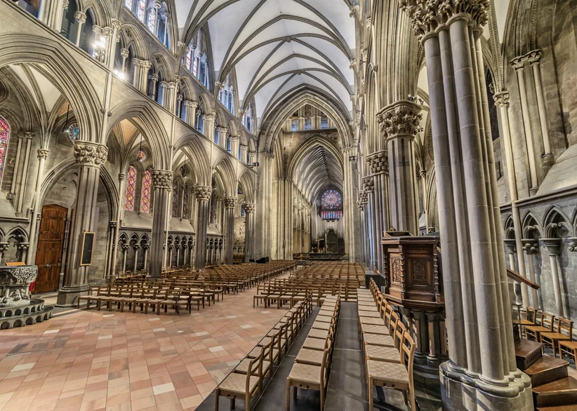 The Gothic nave of Nidaros Cathedral with soaring vaulted ceilings, tall stone columns, rows of wooden pews, and a rose stained-glass window glowing at the far end of the chancel.