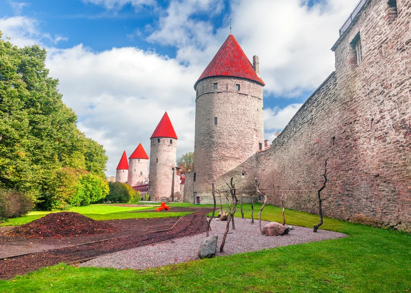 A row of red-roofed medieval defense towers lines Tallinn's stone city wall, overlooking a landscaped garden with sculpted dry branches and gravel beds beneath a blue sky with fluffy clouds.