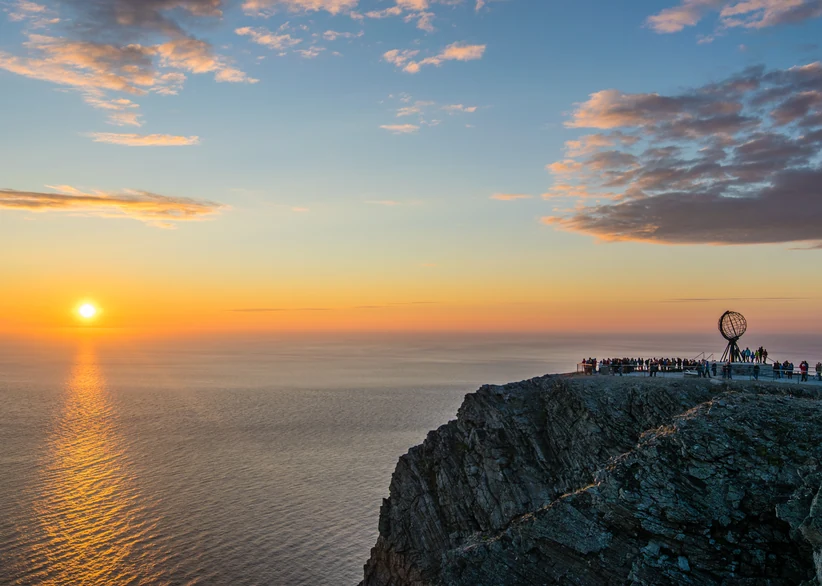 Visitors gather on the clifftop at North Cape around the metal globe monument as the midnight sun sets low over the Arctic Ocean, casting a golden reflection across the water beneath a sky streaked with pink-tinged clouds.