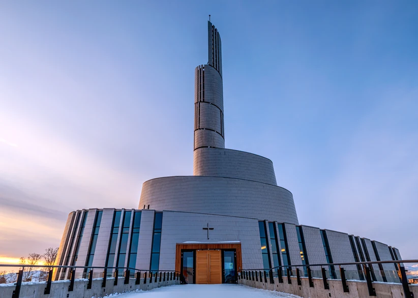The Northern Lights Cathedral's spiraling, titanium-clad tower and curved main structure rise against a soft dusk sky, with a wooden entrance door and cross visible at the base, snow covering the ground.