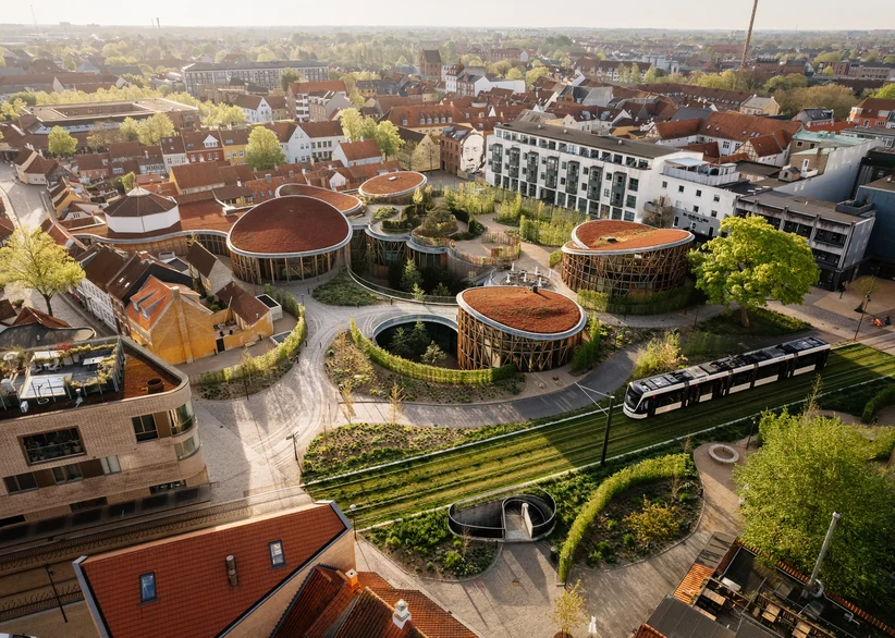 Aerial view of the circular, terracotta-roofed pavilions of H.C. Andersen's House museum set among green gardens in Odense's old town, with a modern tram passing on tracks in the foreground and the red-roofed city skyline beyond.