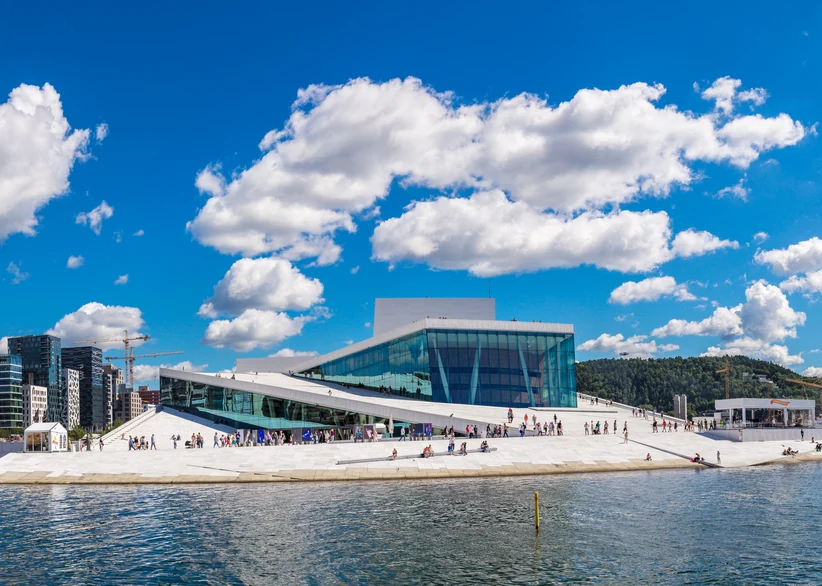 The Oslo Opera House's angular white marble roof slopes down to the waterline, with visitors walking on its sloped surfaces, glass facade reflecting the sky, and city buildings visible in the background under a blue sky with fluffy clouds.