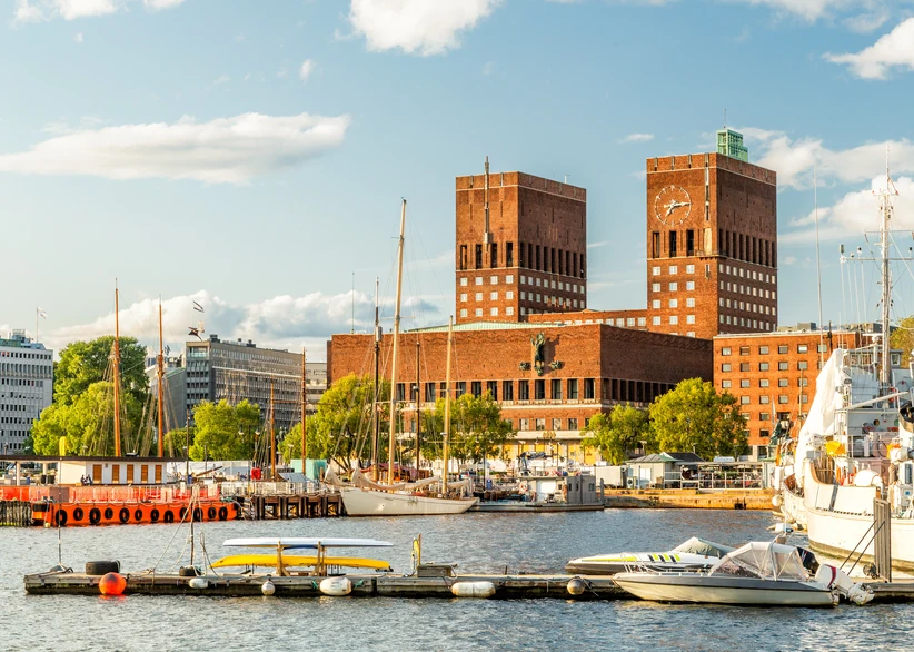 Oslo City Hall's twin brick towers rise above the harbor, with sailboats and motorboats moored along the waterfront in the foreground under a partly cloudy blue sky.
