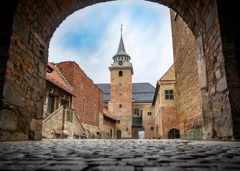 View through a stone archway toward Akershus Fortress's medieval brick tower with its pointed spire and clock, flanked by weathered brick buildings along a cobblestone courtyard.