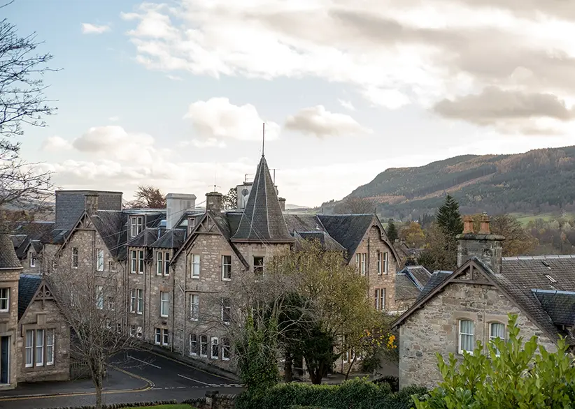 Stone Victorian buildings with slate roofs and a turret in the Highland town of Pitlochry with forested hills and autumn colours in the background, Scotland