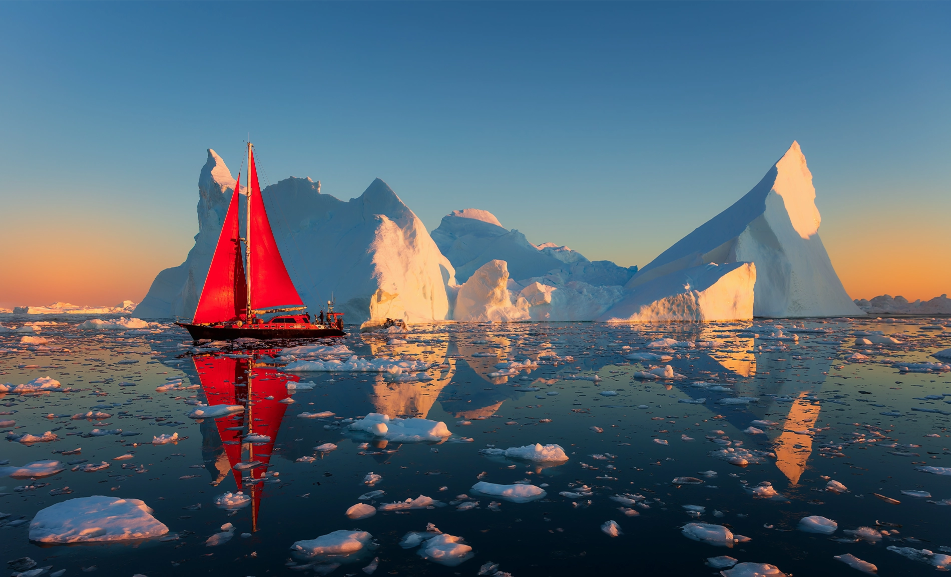 Sailing ship with bright red sails passing towering icebergs on calm, ice-strewn water.