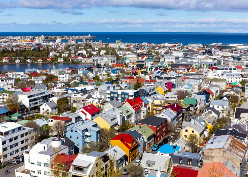 Aerial view of Reykjavik's colorful rooftops in red, blue, yellow, and green, spreading across the cityscape toward the ocean and coastline under a partly cloudy sky.