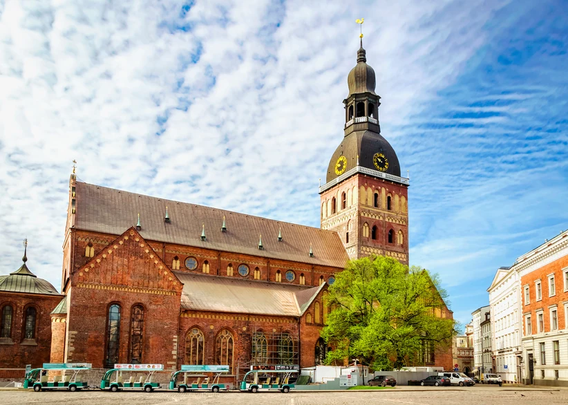Riga Dome Cathedral's red-brick facade and tall black spire with a golden weathervane rise above a cobblestone square, with a green tree, historic buildings, and parked tour vehicles in the foreground.
