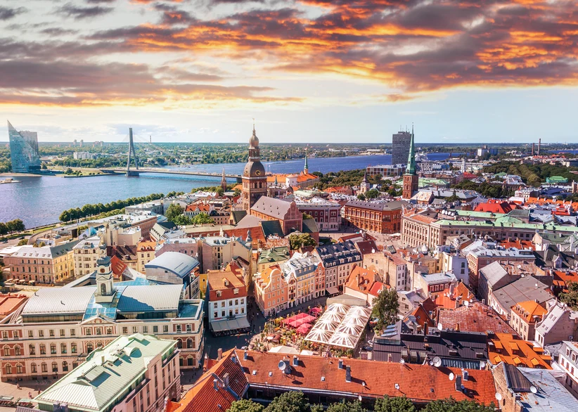 Aerial view of Riga's Old Town at sunset, with Riga Cathedral's tower rising among red-tiled rooftops, the Daugava river and city skyline in the background beneath a dramatic orange and gray sky.