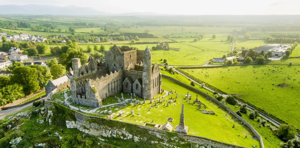 Panoramic aerial view of the Rock of Cashel's medieval cathedral, round tower, and graveyard on a limestone hilltop with the town of Cashel and green Tipperary farmland stretching to distant mountains