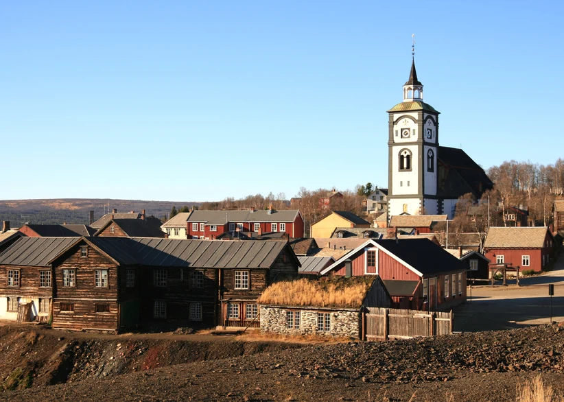 The white church tower of Røros rises above a cluster of dark wooden and red timber houses with turf and slate roofs, set against a backdrop of dark mining slag heaps under a clear blue sky.