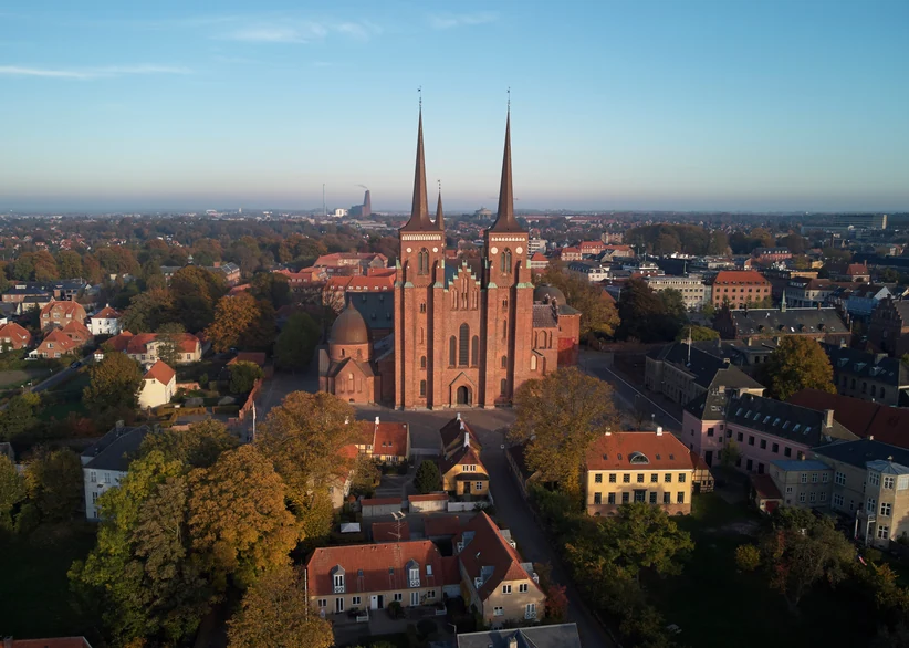 Aerial view at golden hour of Roskilde Cathedral's twin red-brick spires rising above the town's red-roofed houses and autumn trees, with the surrounding cityscape stretching to the horizon.