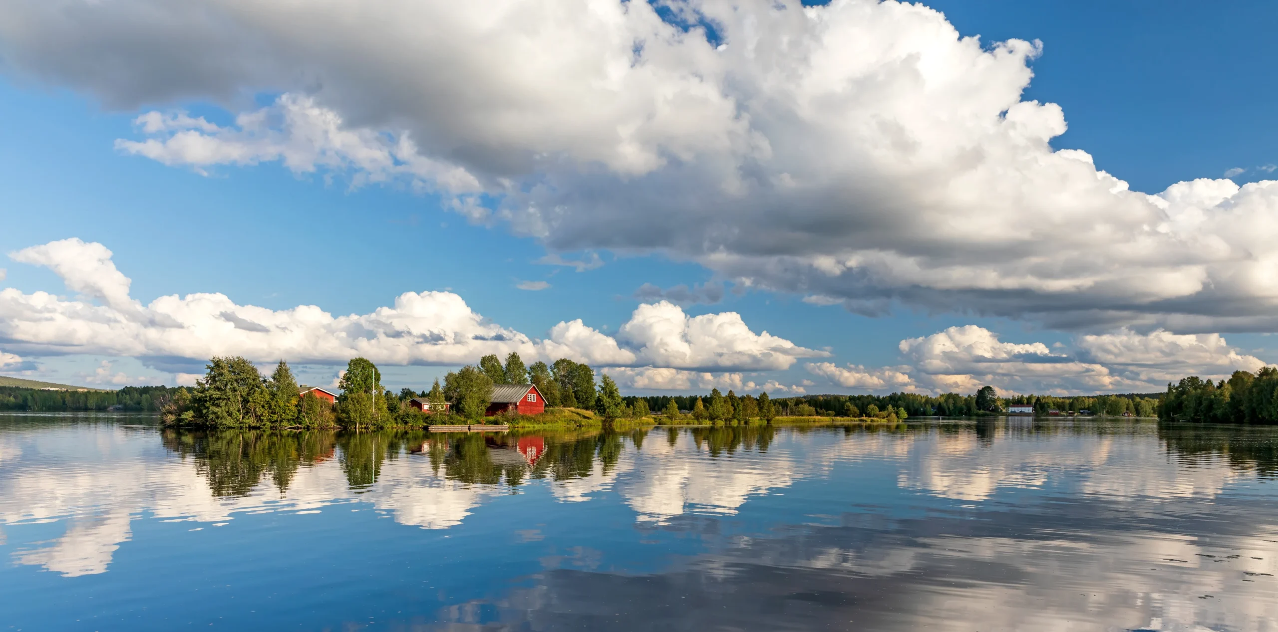 Red wooden cottages on the shore of a lake near Rovaniemi are mirrored perfectly in the still water beneath a dramatic sky of billowing white clouds and blue sky.