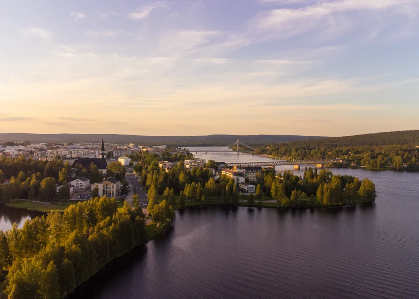 Aerial sunset view of Rovaniemi's church spire and city skyline along a bend in the Kemijoki river, with a bridge crossing the water and dense forested hills stretching to the horizon under a soft golden sky.