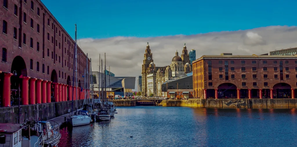 Sailboats moored beside red-columned brick warehouses at Royal Albert Dock, with the Royal Liver Building rising behind under a blue sky.