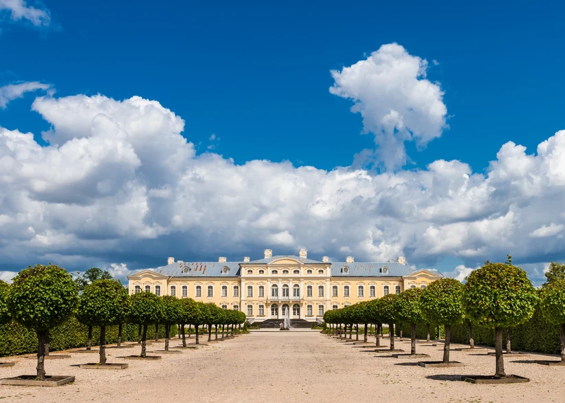 Rundāle Palace's pale yellow baroque facade is framed by symmetrical rows of manicured potted trees lining a gravel path, beneath a bright blue sky with large billowing clouds.