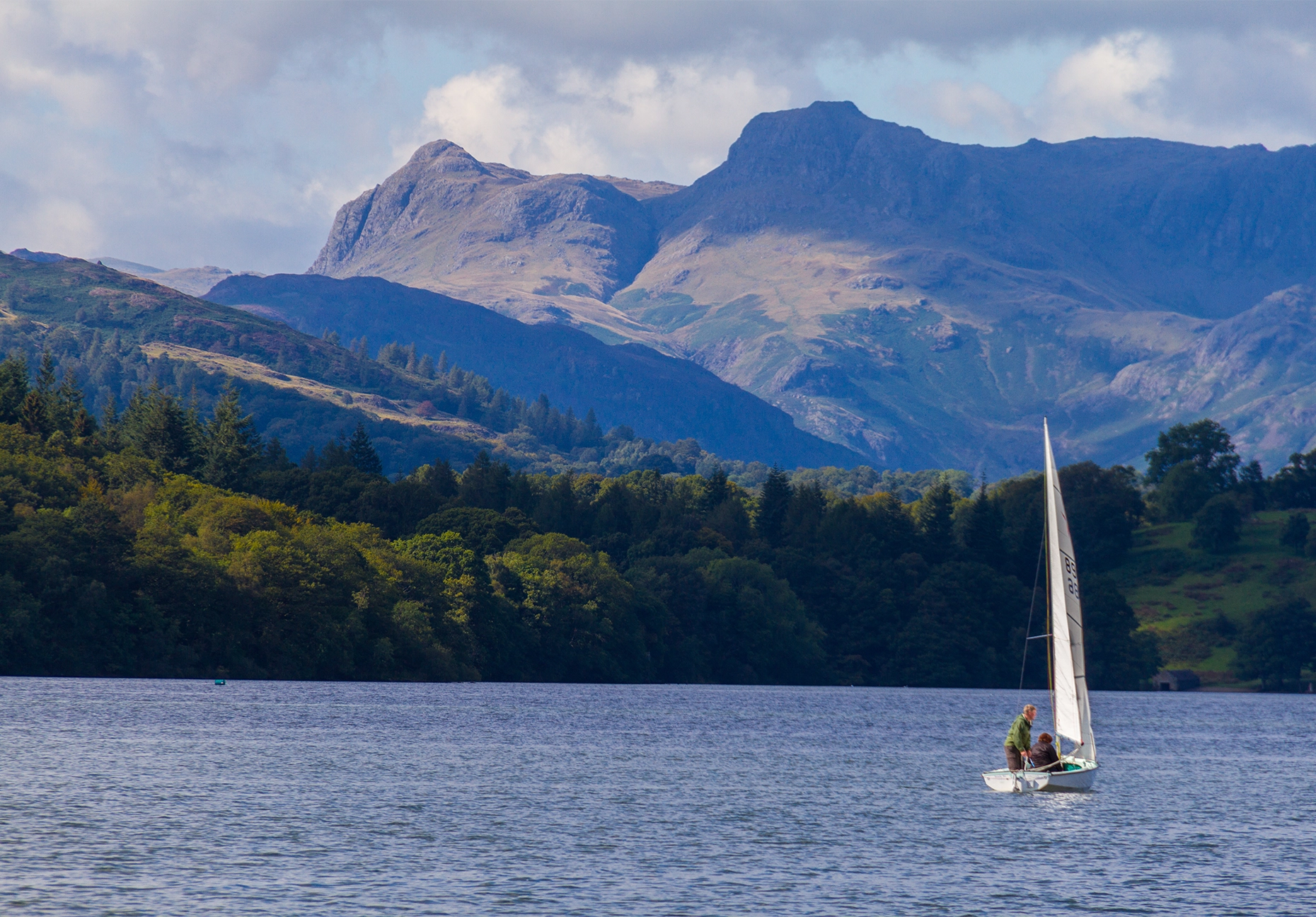 Two people sail a small dinghy on Windermere, with wooded shores and mountains beyond.