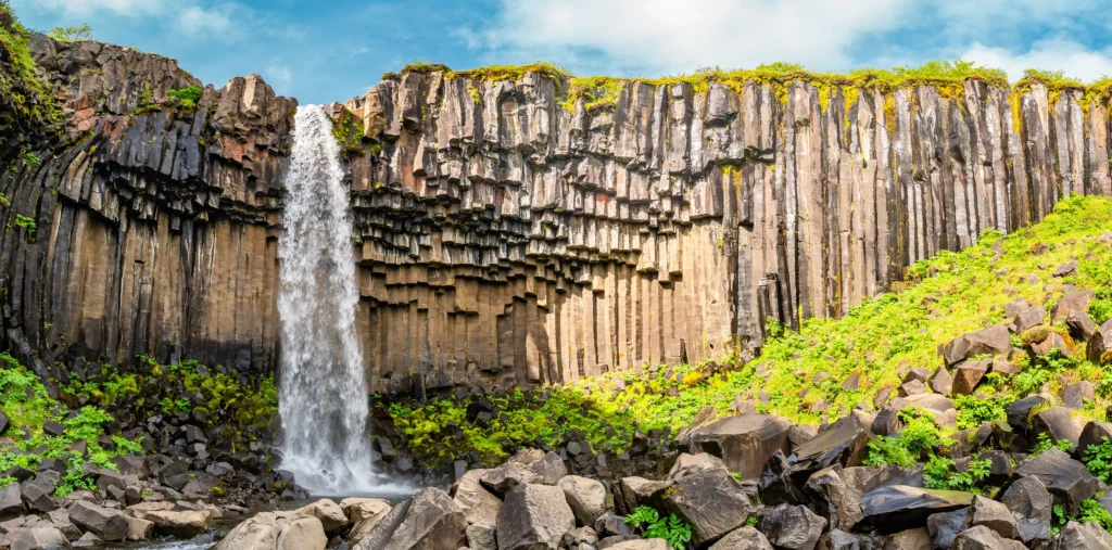 Svartifoss waterfall pours over a cliff framed by dark hexagonal basalt columns, with a moss-covered rock field and stream at its base under a partly cloudy sky.