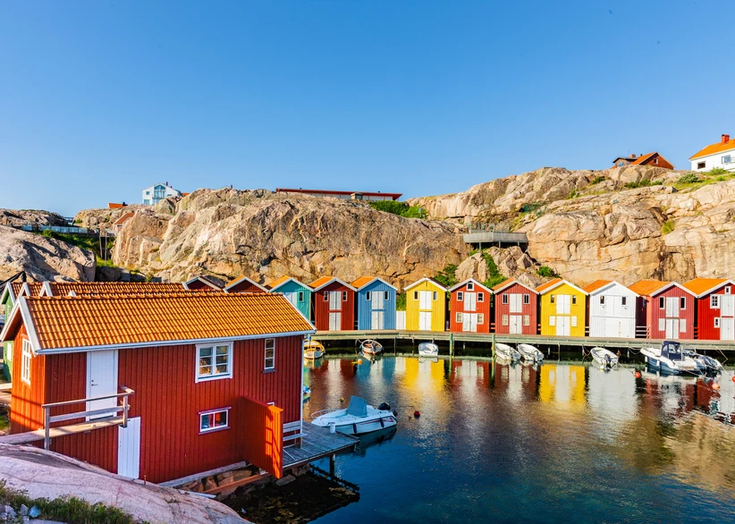 A row of colorful wooden boathouses in red, yellow, and blue line a marina in Smögen, with small boats moored along the dock and bare granite rocks rising behind under a clear blue sky.