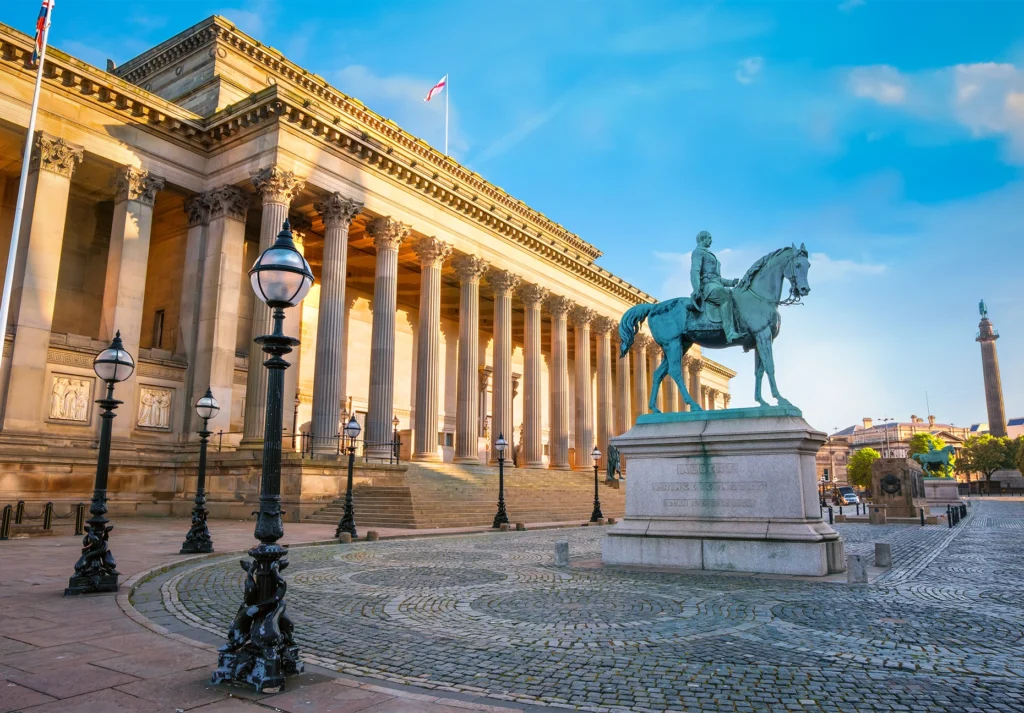 A neoclassical columned hall beside a bronze equestrian statue on a cobbled square in golden light.