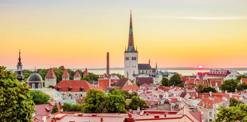 St. Olaf's Church's tall spire rises above Tallinn's red-tiled rooftops and medieval towers at sunset, with the sea visible in the distance beneath a soft orange and yellow sky.