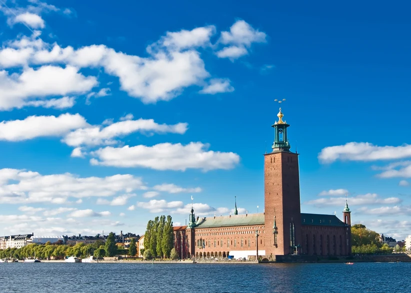 Stockholm City Hall's tall red-brick tower with its golden spire rises above the waterfront, seen across a blue harbor under a sky full of white clouds.