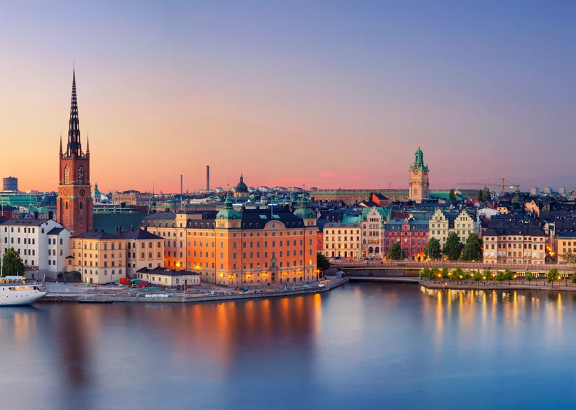 Riddarholmen's spired church and Gamla Stan's colorful waterfront buildings glow at sunset in Stockholm, reflected in the still harbor water beneath a pastel pink and blue sky.