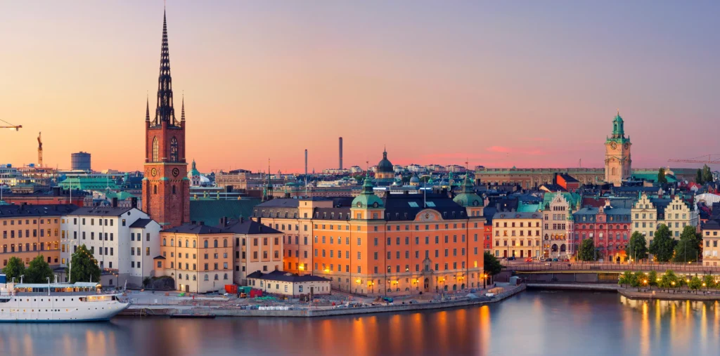 Stockholm's Gamla Stan old town glows at sunset, with the Riddarholm Church's black spire and the colorful waterfront buildings reflected in the still harbor water, a moored ferry in the foreground, and a pink sky over the city skyline.