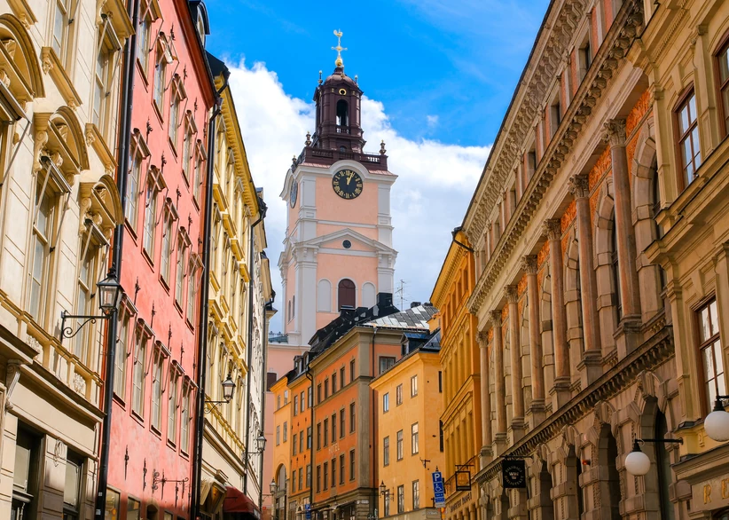 Storkyrkan's ornate clock tower rises above a narrow cobblestone street lined with colorful pink, orange, and cream buildings in Stockholm's Gamla Stan old town, under a blue sky with scattered clouds.