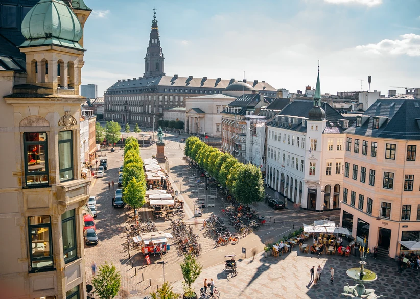 Elevated view of Højbro Plads square in central Copenhagen, lined with tree-shaded cafes, parked bicycles, and outdoor market stalls, with Christiansborg Palace's spire rising in the background under a sunny sky.