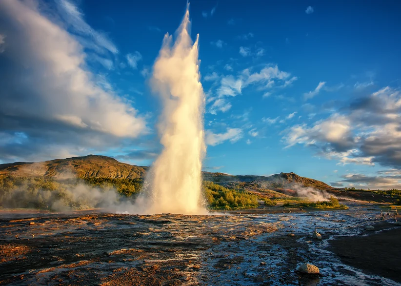 Strokkur geyser erupts a tall plume of steaming water into a bright blue sky filled with clouds, surrounded by a geothermal field of rocky terraces and distant hills.