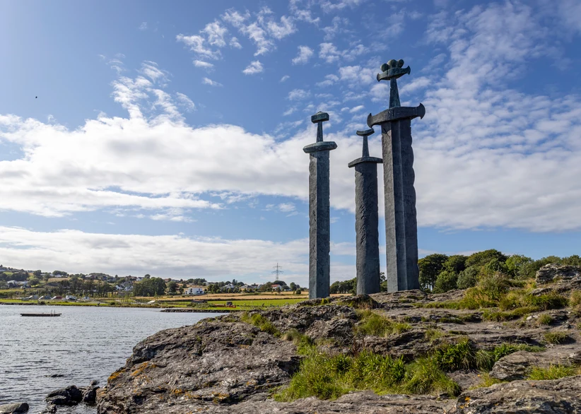 Three giant bronze and stone swords of the Sverd i Fjell monument stand planted in a rocky coastal outcrop overlooking a fjord, with a grassy shoreline and distant hills under a blue sky with scattered clouds.