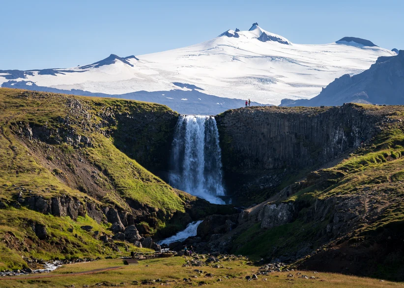 Svöðufoss waterfall cascades over a basalt cliff into a rocky river valley, with two hikers standing at the edge and the snow-capped Snæfellsjökull glacier volcano rising in the background.