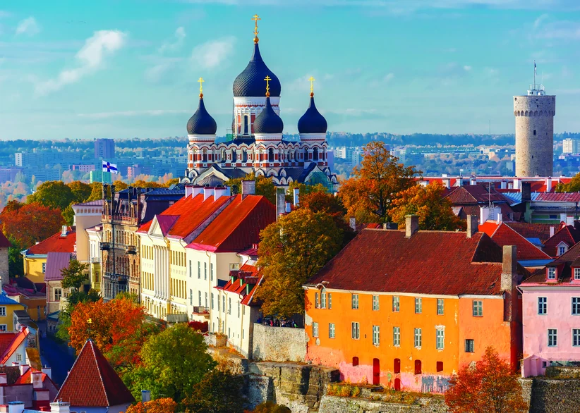Alexander Nevsky Cathedral's dark onion domes rise above Toompea Hill's autumn-colored trees and colorful red-roofed buildings, with the medieval Pikk Hermann tower visible in the background under a blue sky.