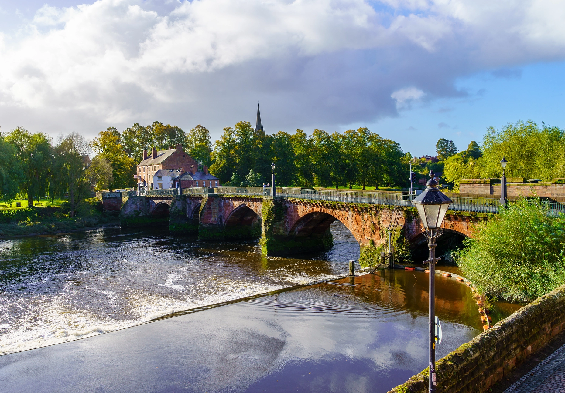 A red sandstone arched bridge crosses a river beside a weir, with a church spire rising above autumn trees.