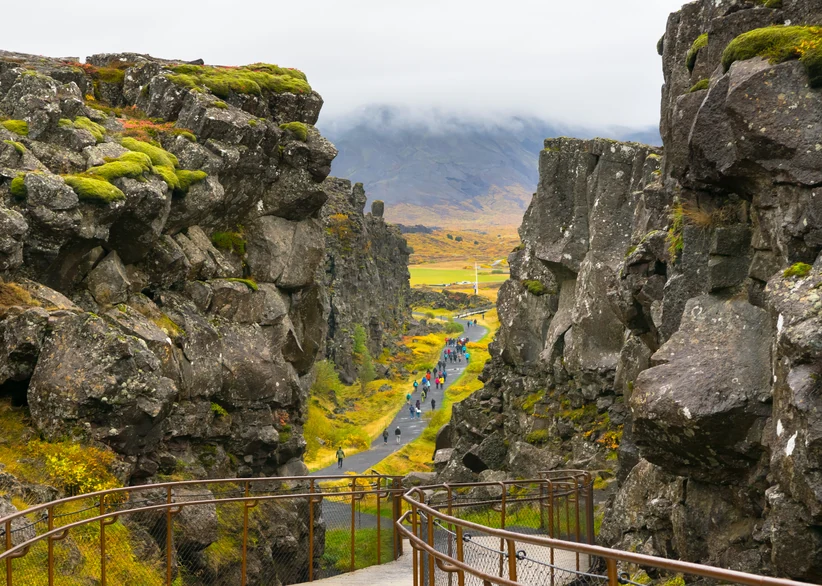 Visitors walk along a wooden boardwalk through the rocky, moss-covered rift between Iceland's tectonic plates at Þingvellir National Park, with fog-shrouded mountains and a golden valley visible in the distance.